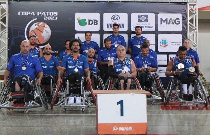 Vários homens em cadeiras de rodas que formam um time de Basquete em cadeira de rodas, posam para uma foto de grupo, destacando as medalhas que ganharam.