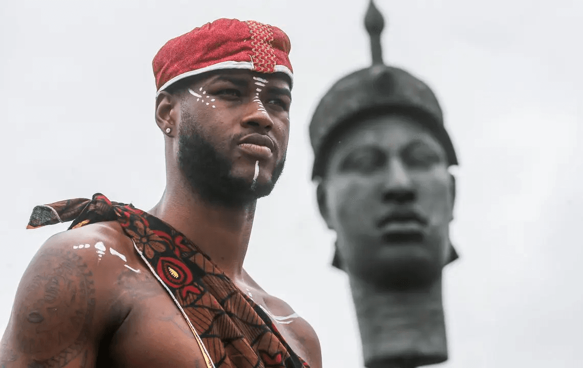 Imagem de um homem com vestimentas tradicionais africanas, em destaque, com pintura corporal, contemplando uma estátua. Cenário cultural significativo.