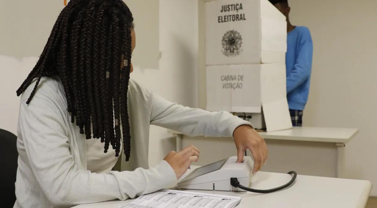 Mulher com dreadlocks votando em uma cabine eleitoral, demonstrando participação cívica em um local de votação.