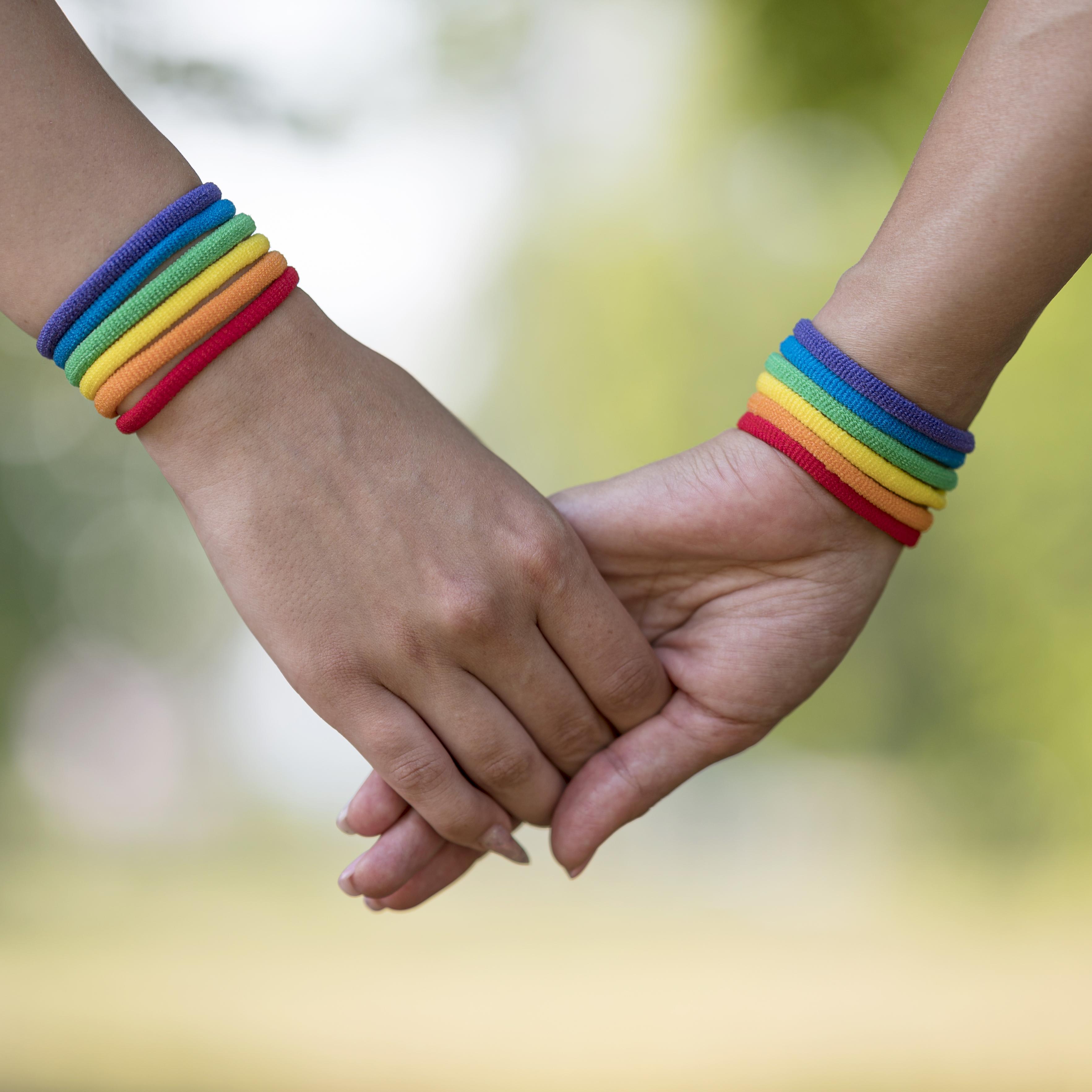 Duas mãos dadas com pulseiras das cores da bandeira LGBTQUIAP+, representando uma relação de afeto.