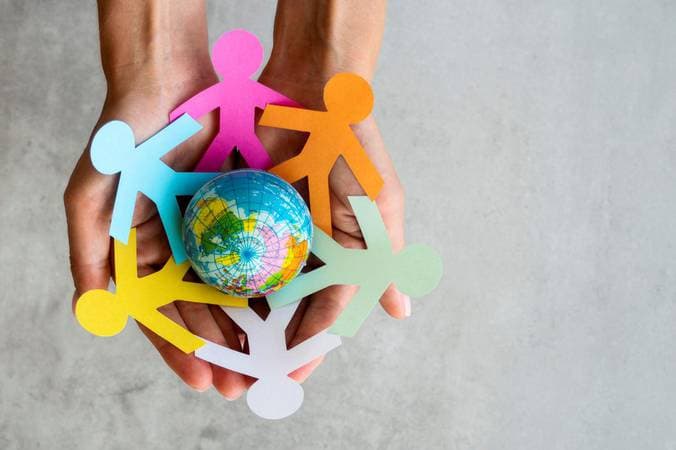 Mãos segurando um pequeno globo terrestre cercado por bonecos de papel colorido, representando diversidade e direitos humanos.