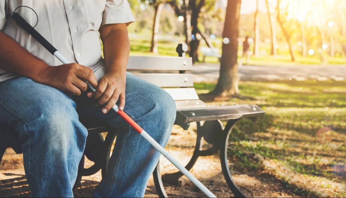 Pessoa sentada em um banco de parque, segurando uma bengala branca e vermelha (típica para deficientes visuais), com o sol brilhando ao fundo.