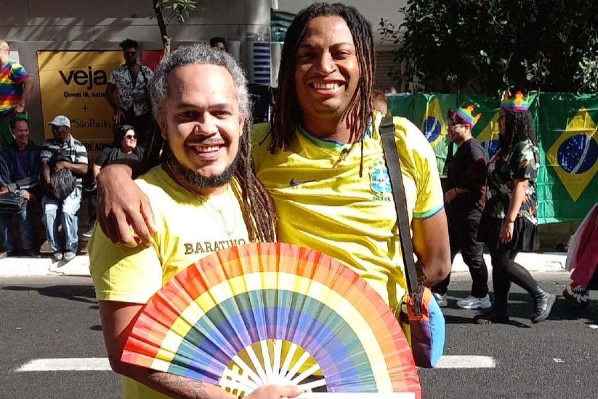 A imagem mostra dois homens sorrindo e abraçados em um evento ao ar livre. Ambos estão usando camisetas amarelas da seleção brasileira de futebol. Um dos homens segura um leque com as cores do arco-íris, símbolo do orgulho LGBTQ+. Ao fundo, é possível ver outras pessoas, algumas também com acessórios coloridos e bandeiras do Brasil, indicando que a foto foi tirada durante uma parada LGBTQ+ ou um evento similar de celebração e apoio à diversidade.